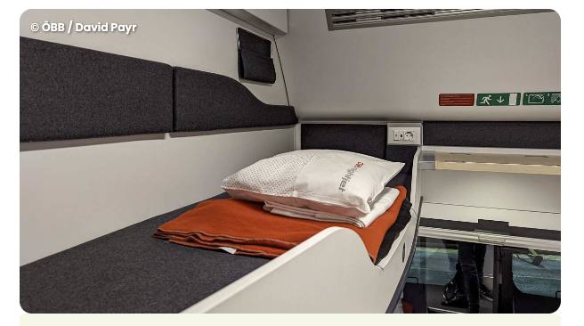 Einzelabteil im neuen ÖBB-Nightjet LUNA Wagen mit frisch bezogenem Bett, orangefarbener Decke, Nightjet-Kissen und Steckdose an der Kabinenwand.